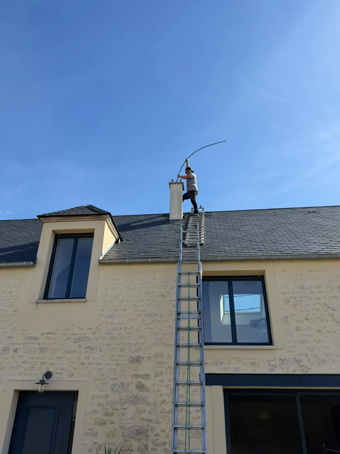 Ramoneur sur le toit d'une maison en pierre, canne de ramonage engagée dans la souche de cheminée, dans le Loiret